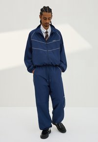 Moški s spletenimi lasmi, oblečen v mornarsko modro športno obleko preko bele srajce in črtaste kravate, stoji z rokami v žepih in gleda navzdol.