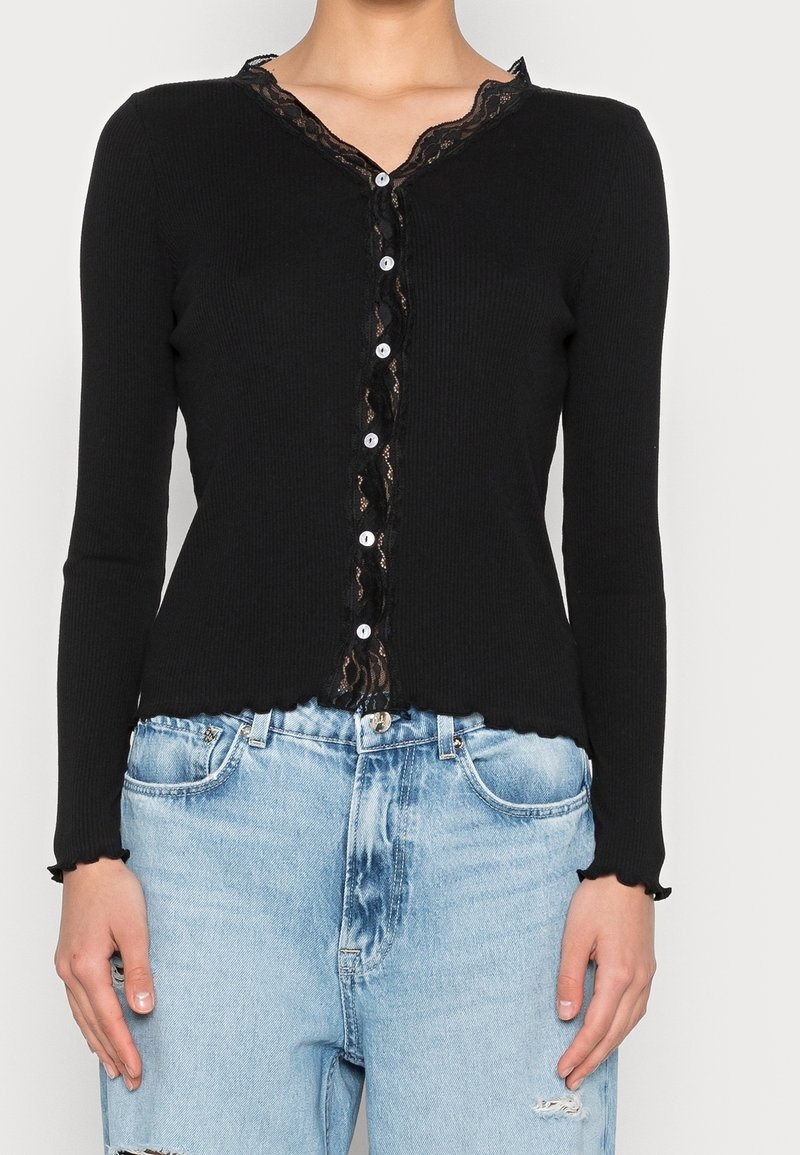Cardigan nero a coste con maniche lunghe, bordino in pizzo ondulato lungo il scollo a V e sei bottoni bianchi sul davanti. Indossato sopra jeans azzurri chiari.
