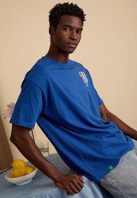 Koninklijk blauwe katoenen t-shirt met korte mouwen, voorzien van een grafische print in oranje aan de linkerkant. Casual pasvorm, gecombineerd met lichtblauwe jeans.