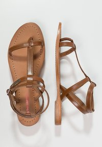 Bruine leren sandalen met platte zolen. Bevat meerdere kruissgewoven banden en een ronde neus. Zijaanzicht toont een lage hak en stiksels.