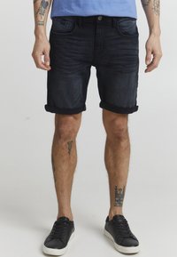 Dongerishorts i mørk vask med oppbrettede kanter, fem lommer og subtil falming. Brukt med svarte sneakers på en lys bakgrunn.