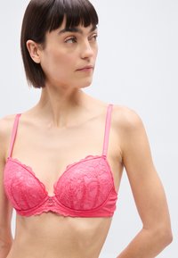 Γυναίκα με κοντά σκούρα μαλλιά, φοράει ένα ροζ δαντελωτό σουτιέν, κοιτάζοντας προς τα αριστερά σε φόντο απλό φωτεινό.