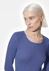 Jeune femme aux cheveux blonds clairs portant un t-shirt bleu côtelé à manches longues ajusté, regardant vers le bas et sur le côté sur un fond blanc.