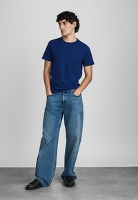 Νέος άντρας στέκεται με τα χέρια στις τσέπες, ντυμένος με μπλε σορτσάκι, φαρδιά τζιν παντελόνια και μαύρα παπούτσια, κοιτάζοντας αριστερά.