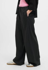 Pantalons noirs à jambes larges avec une taille à cordon, poches latérales et une texture lisse. Associés à des baskets argentées et un haut rose.