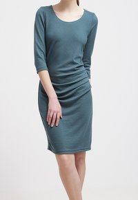 Robe en jersey couleur bleu sarcelle avec manches trois-quarts, encolure dégagée, et côtés froncés. Silhouette ajustée, longueur genou, texture lisse.