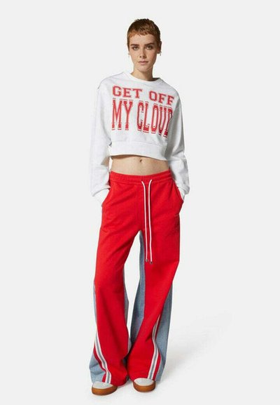 Sweatshirt bianco tagliato con lettering rosso e pantaloni felpa rossi con pannelli grigi e strisce bianche, abbinati a scarpe beige.