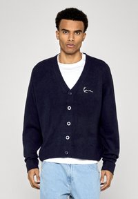 Námořnický pletený cardigan s véčkovým výstřihem, pěti předními knoflíky, žebrovanými manžetami a malým bílým logem na levém boku hrudi. Nosený přes bílou košili.