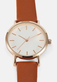 Montre ronde avec un cadran blanc, des index en or rose et un boîtier en laiton. Équipée d'un bracelet en cuir brun à la texture lisse.