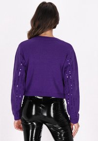 Pulover scurt violet, cu un corp din tricot texturat și mâneci lungi, decorate cu rânduri verticale de paiete. Asortat cu colanți negri strălucitori.