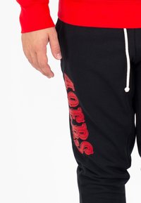 Pantaloni della tuta neri con testo grafico rosso "40RS" lungo il lato, dotati di vita con coulisse e texture liscia. Abbinati a una maglietta a maniche lunghe rossa.