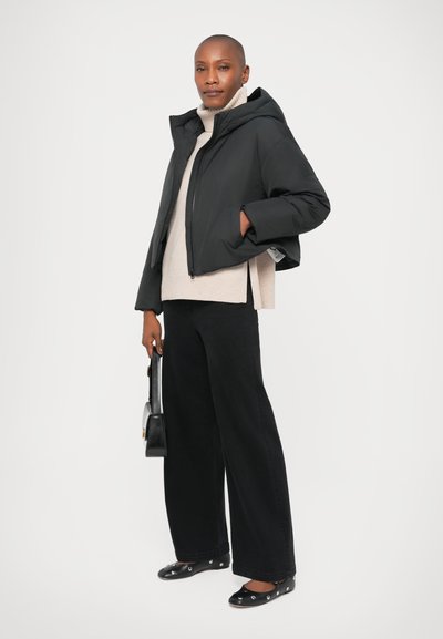 Chaqueta acolchada negra corta con capucha, suéter de cuello alto beige, jeans negros de pierna ancha, bolso negro y zapatos planos con acentos metálicos.