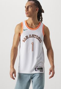 Camiseta de baloncesto blanca con ribetes naranjas, que presenta "SAN ANTONIO" y el número "1" en marrón. Incluye el logo de Nike y detalles de la etiqueta de la NBA.