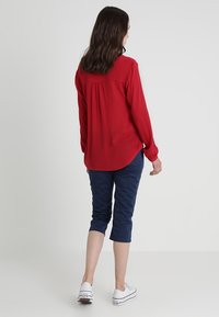 Blusa rossa a maniche lunghe con schiena plissettata, abbinata a pantaloni navy a 3/4. Il modello indossa sneakers bianche. Design di outfit semplice e casual.