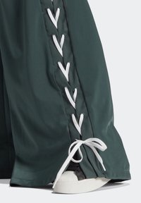 Zelené saténové kalhoty s bílými krajkovými detaily na straně a mašlemi na lemu, které ukazují tenisku vykukující zespodu.