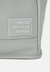 Esquina de una bolsa gris con una etiqueta blanca que dice "REUTILIZAR RECICLAR REPETIR" en letras mayúsculas.