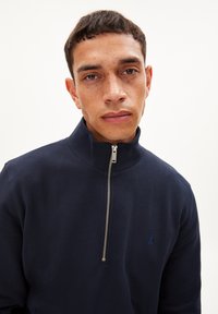 Navyblauwe fleece pullover met een hoge kraag en een zilveren rits. Heeft een klein, subtiel logo op de linkerbovenborst. Glad textuur.