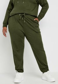 Pantaloni jogger a coste verde oliva con cintura regolabile, tasche laterali, gambe affusolate, indossati con sneakers bianche. Dettaglio del tessuto testurizzato visibile.