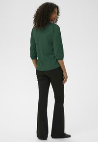 Bluză verde cu fuste încrețite, cu mâneci trei sferturi, asortată cu pantaloni negri evazați. Bluza are un guler rotund și o margine ușor curbată.
