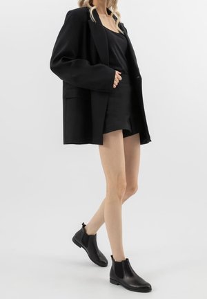Kobieta ubrana w czarną oversize marynarkę, czarny top, czarne krótkie spodenki i czarne botki, stojąca na tle jednolitego jasnego tła.