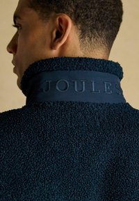 Mies, joka katsoo sivulle, pukeutuneena tummansiniseen tekstiilipintaiseen fleecetakkiin, jossa on "JOULES" brodeerattu nousseeseen kaulukseen.