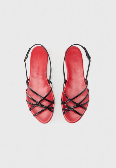 Κόκκινα δερμάτινα σανδάλια με μαύρους σταυρωτούς ιμάντες, ανοιχτό σχέδιο δακτύλου και κούμπωμα με πόρπη. Λείο υφή με επίπεδη σόλα.