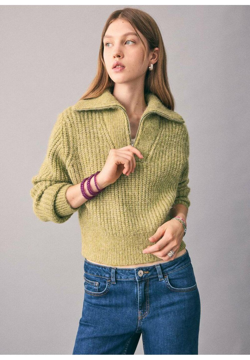 Pulover tricotat de culoare verde deschis, cu guler larg și fermoar, asortat cu blugi de culoare albastru închis. Modelul poartă brățări violete și cercei din argint.