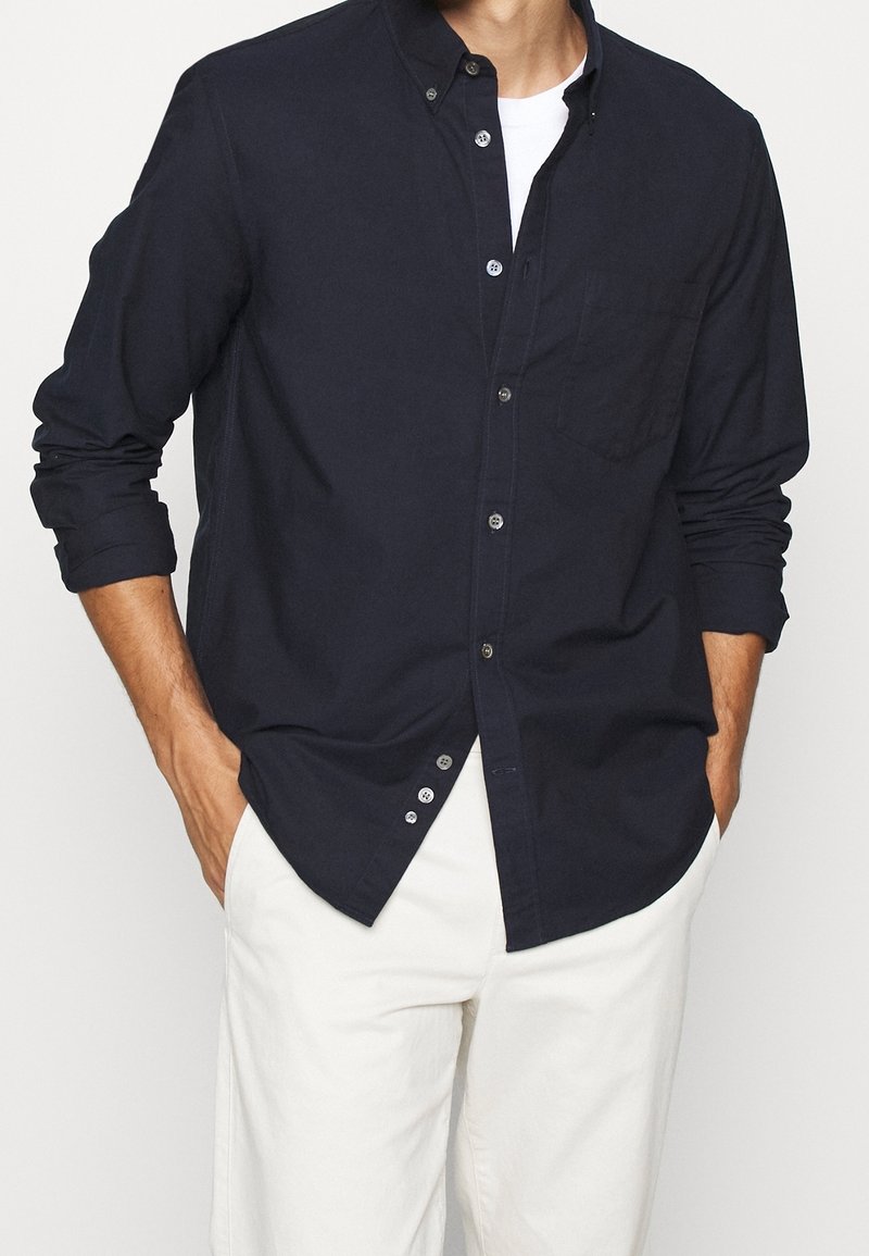 Homme portant une chemise bleu marine à boutons par-dessus un t-shirt blanc avec les manches retroussées et un pantalon blanc, les mains dans les poches.
