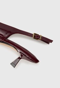 Burgundy patento nahka hedelmäkengät, joissa on terävä kärki, säädettävä nilkkaremm ja pieni lohkokorko. Kullattu solki yksityiskohtana remmissä.