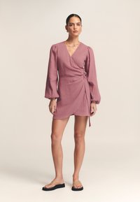 Obleka z zavezovanjem v svetlo roza barvi, izdelana iz teksturirane tkanine. Ima dolge, puhaste rokave in detajl zavezovanja v pasu. Videz dopolnjujejo ravne sandale.