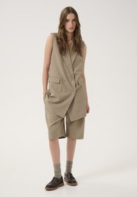 Beige double-breasted vest with large buttons, paired with loose shorts. Brown lace-up shoes and gray socks complete the outfit.