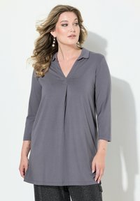 Tunic grigio a maniche lunghe con scollo a V, dettaglio plissettato sul davanti e tessuto morbido. Abbinato a pantaloni neri testurizzati, creando un look a strati.