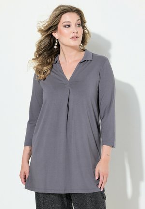Grijze tuniek met lange mouwen en V-hals, met geplooide details aan de voorkant en een zachte stof. Gestyled met gestructureerde zwarte broeken, wat een gelaagde look creëert.