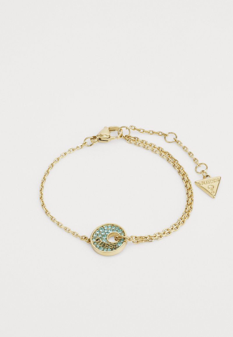 Bracelet chaîne en or avec un charm rond incrusté de petites pierres bleues et le nom de la marque, plus une petite étiquette triangulaire de la marque sur le fermoir.