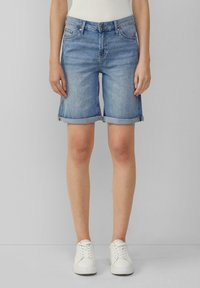 Pantalones cortos de mezclilla azul claro con dobladillos enrollados, cinco bolsillos y cierre frontal con botón. Llevados con zapatillas blancas y una blusa ajustada.
