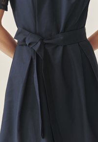 Tumesinine kleit, millel on keha järgiv ülakeha, lühikesed pitsvarrukad ja vööga seotud talje; siledast kangast seeliku paeltes lisanduva detailiga.