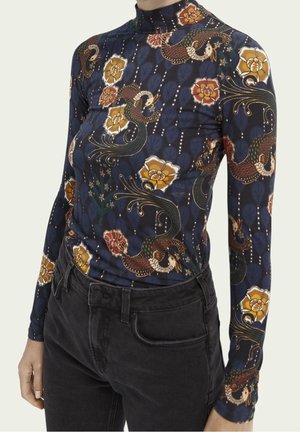 Femme portant un haut bleu marine ajusté à manches longues avec des motifs floraux et d'oiseaux, rentré dans un jean noir.