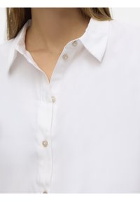 Gros plan d'une personne portant une chemise blanche à col avec des boutons beige, montrant la partie supérieure de la poitrine et la zone du cou.