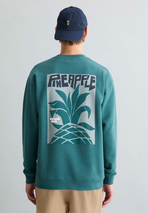 Personne portant un sweat-shirt sarcelle avec un grand graphique stylisé d'ananas et le texte "PINEAPPLE" au dos, ainsi qu'une casquette marine avec un petit emblème d'ananas.