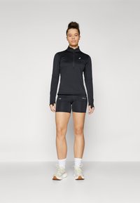 Donna in piedi che indossa un top atletico nero a maniche lunghe con mezzo zip, pantaloni corti neri aderenti, calze bianche e scarpe da corsa beige su sfondo neutro.