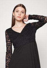 Vestido preto com mangas longas de renda transparente. Design em V com corpete de renda texturizada, que transita para uma saia lisa. Sem padrões ou acessórios.
