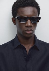 Black and tortoiseshell sunglasses with square frames, dark lenses, and subtle hardware details, worn with a dark blue button-up shirt.