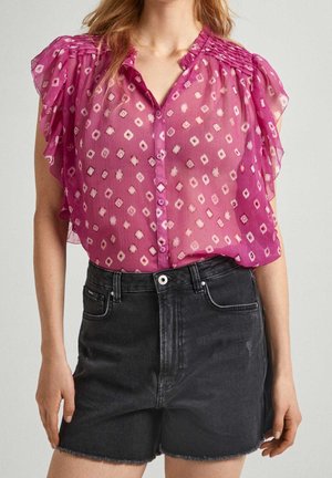 Vrouw draagt een doorschijnende roze blouse met patroon en ruches aan de mouwen, ingestopt in een hooggesneden zwarte spijkershort.