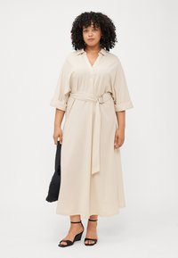 A woman with curly hair wears a beige belted dress and black wedge sandals, holding a textured black bag against a plain background.