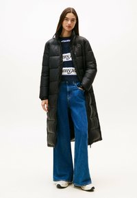 Langer schwarzer Daunenmantel mit hohem Kragen, getragen über einem marineblauen Pullover mit weißer Schrift und weiten blauen Jeans.