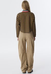 Camicia a quadri gialla e blu scuro, con colletto a strisce rosse, verdi e blu; abbinata a pantaloni beige a gamba larga con tasche laterali.
