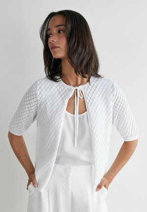 Kobieta o średniej długości brązowych włosach nosi biały, teksturowany kardigan z krótkim rękawem, zawiązany na szyi, na bezrękawniku i białych spodniach.