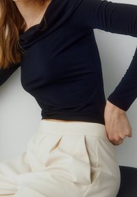 Femme portant un haut noir ajusté à manches longues et un pantalon crème plissé taille haute, assise devant un fond clair uni.