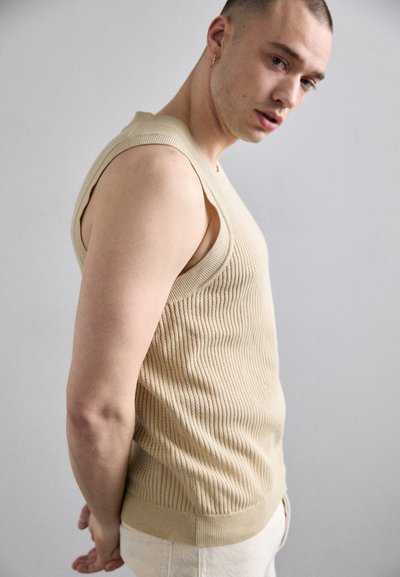 Top básico de canalé beige con amplios huecos para los brazos, que presenta un diseño de punto texturizado. El dobladillo inferior está rematado con una banda sólida y ligeramente más gruesa.