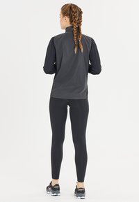 Schwarze Sportjacke mit eng anliegenden Ärmeln, kombiniert mit schwarzen Leggings und Laufschuhen. Der Stoff wirkt glatt und leicht.